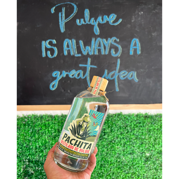 Una mano sosteniendo una botella de destilado de pulque con una frase en el fondo que dice Pulque is always a great idea